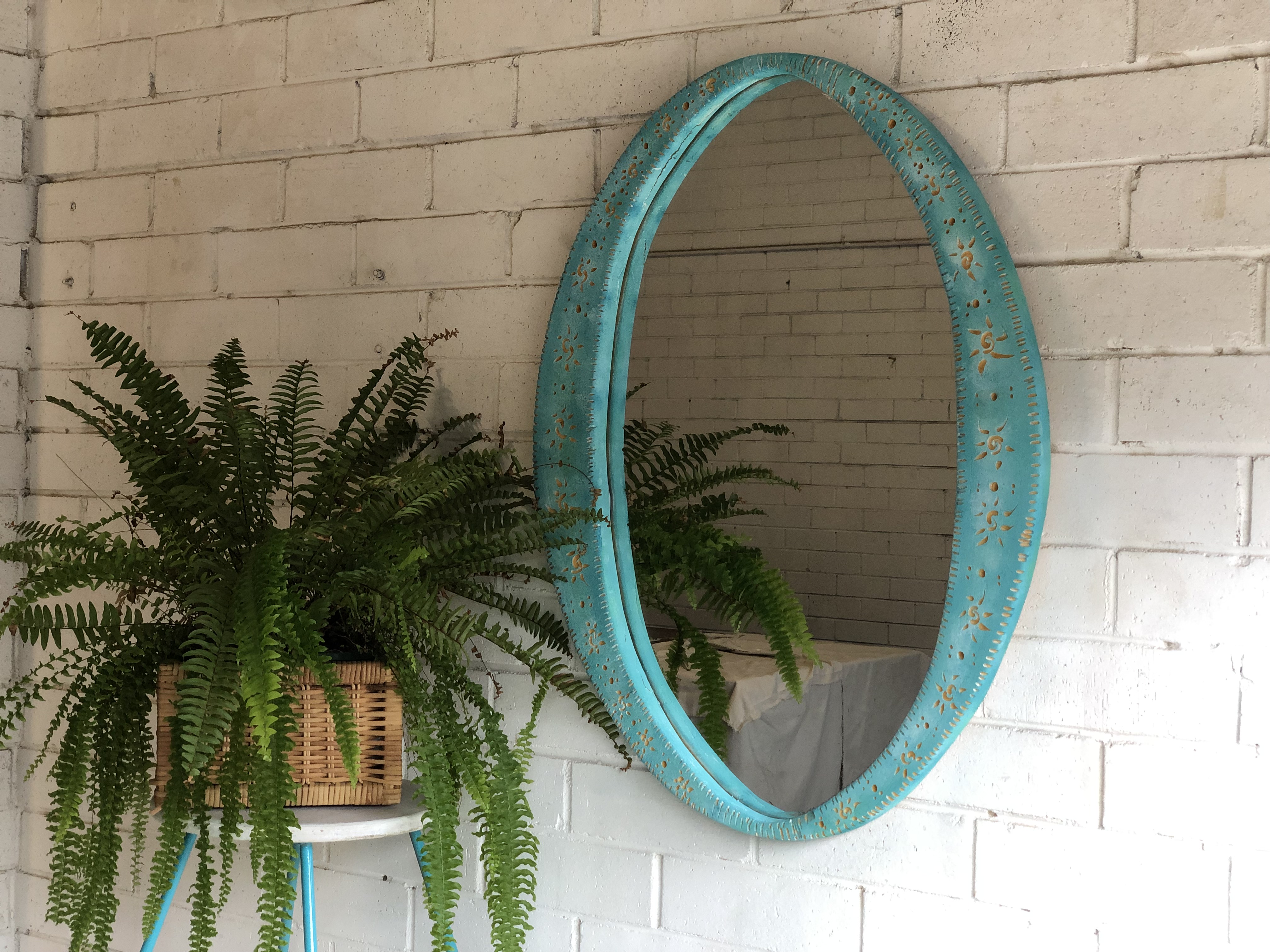 On a white, brick wall hangs a mirror with an ornate blue frame. On the left a fern sits on top of a stool