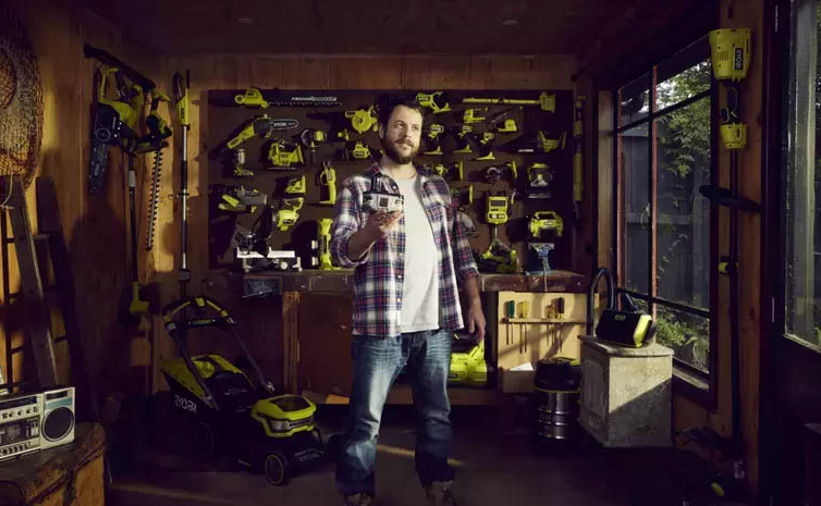 A man holds a 18V battery in a shed filled with RYOBI tools